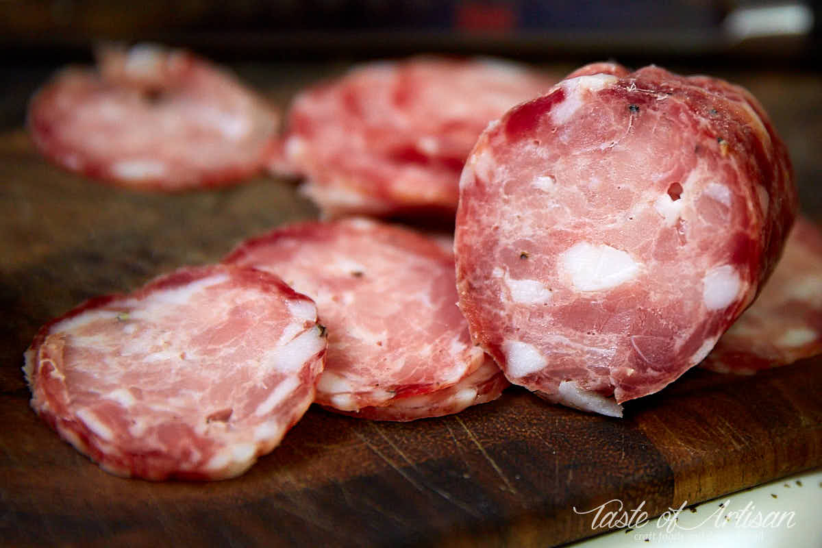 Slices of sopressata on foreground with uncut sopressata on background on a cutting board.