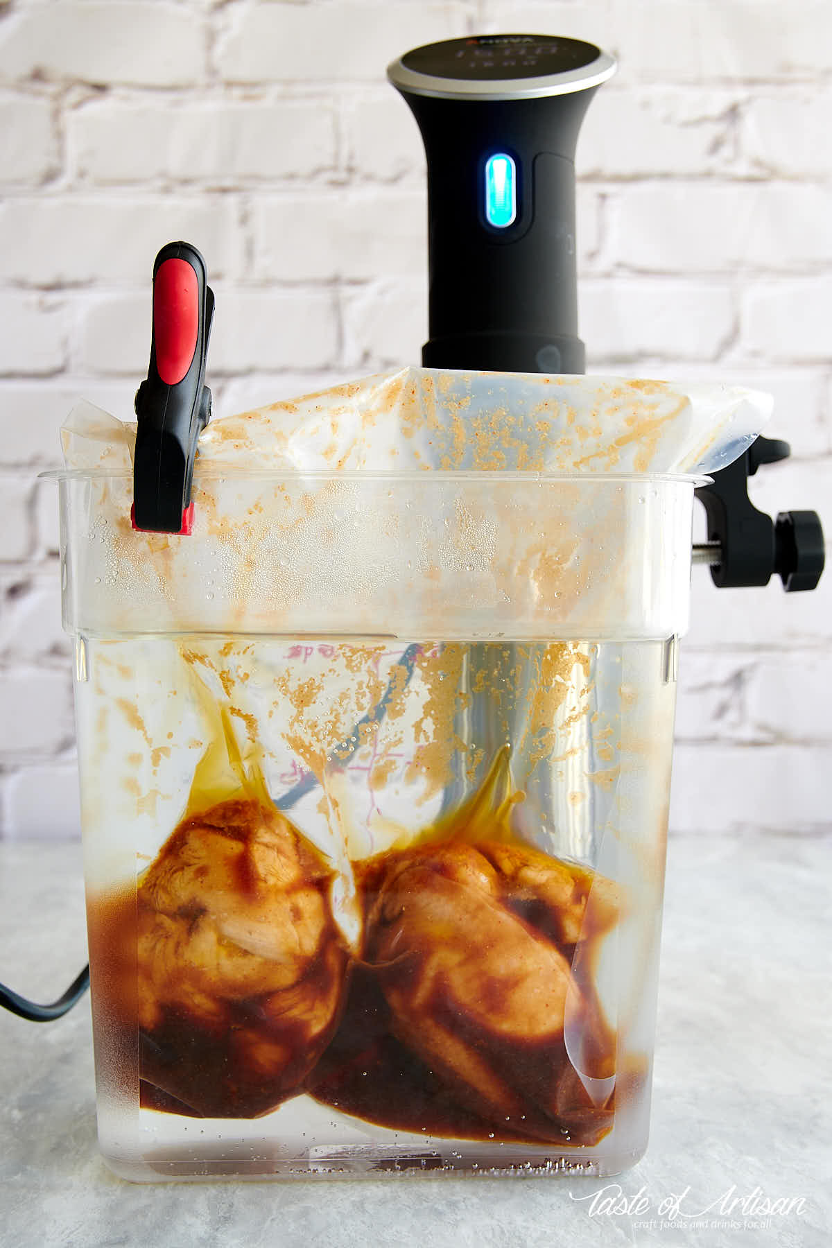 Two chicken breasts in a vacuum sealed bag with sauce cooking inside a clear container filled with water and with a sous vide circulator.