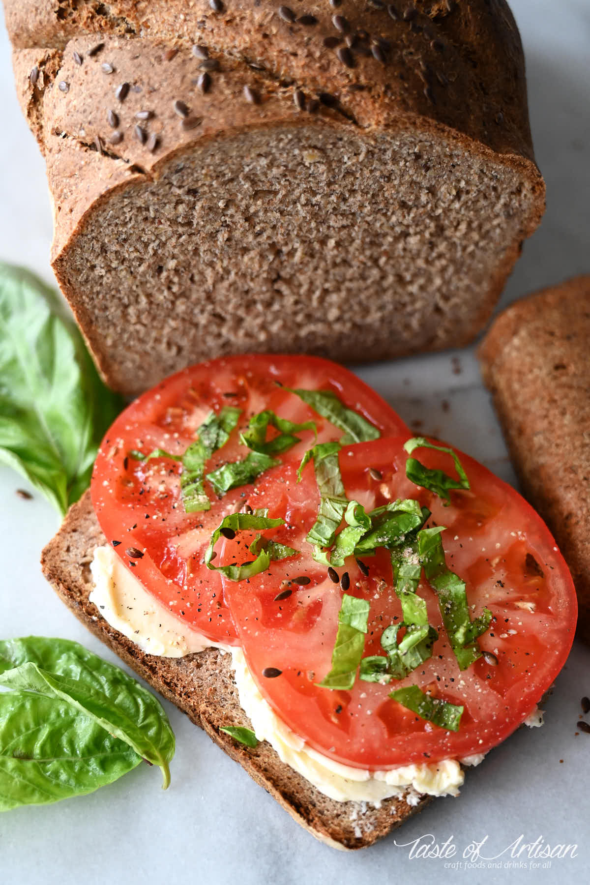 A slice of flaxseed bread with butter and slices of tomatoes and chopped basil on top.