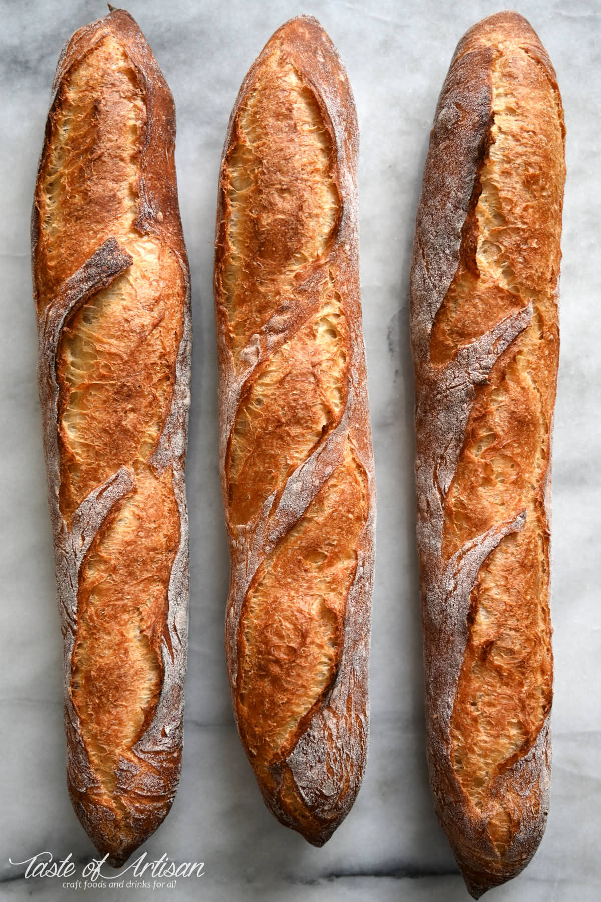 Three golden brown homemade baguettes on a marble table.