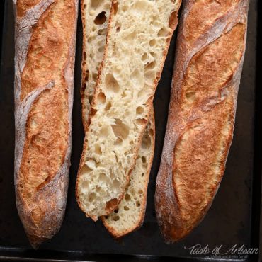 Three homemade French baguettes on a baking sheet, one cut open in half showing airy crumb.