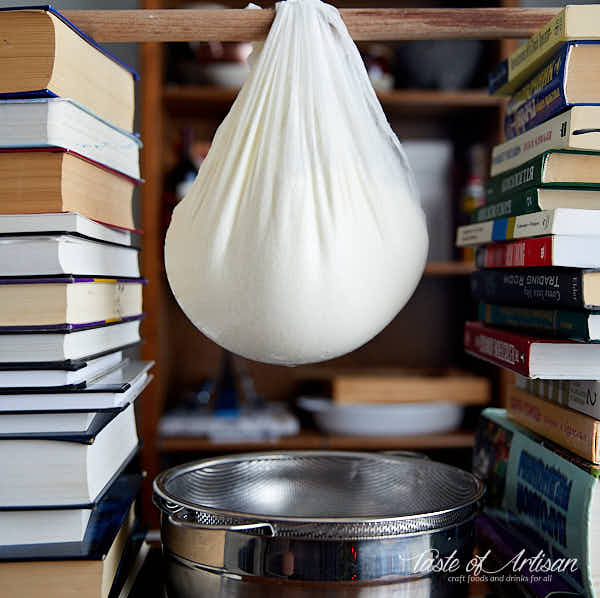Cream cheese curd draining in a muslin hanging from a stick over a bowl.