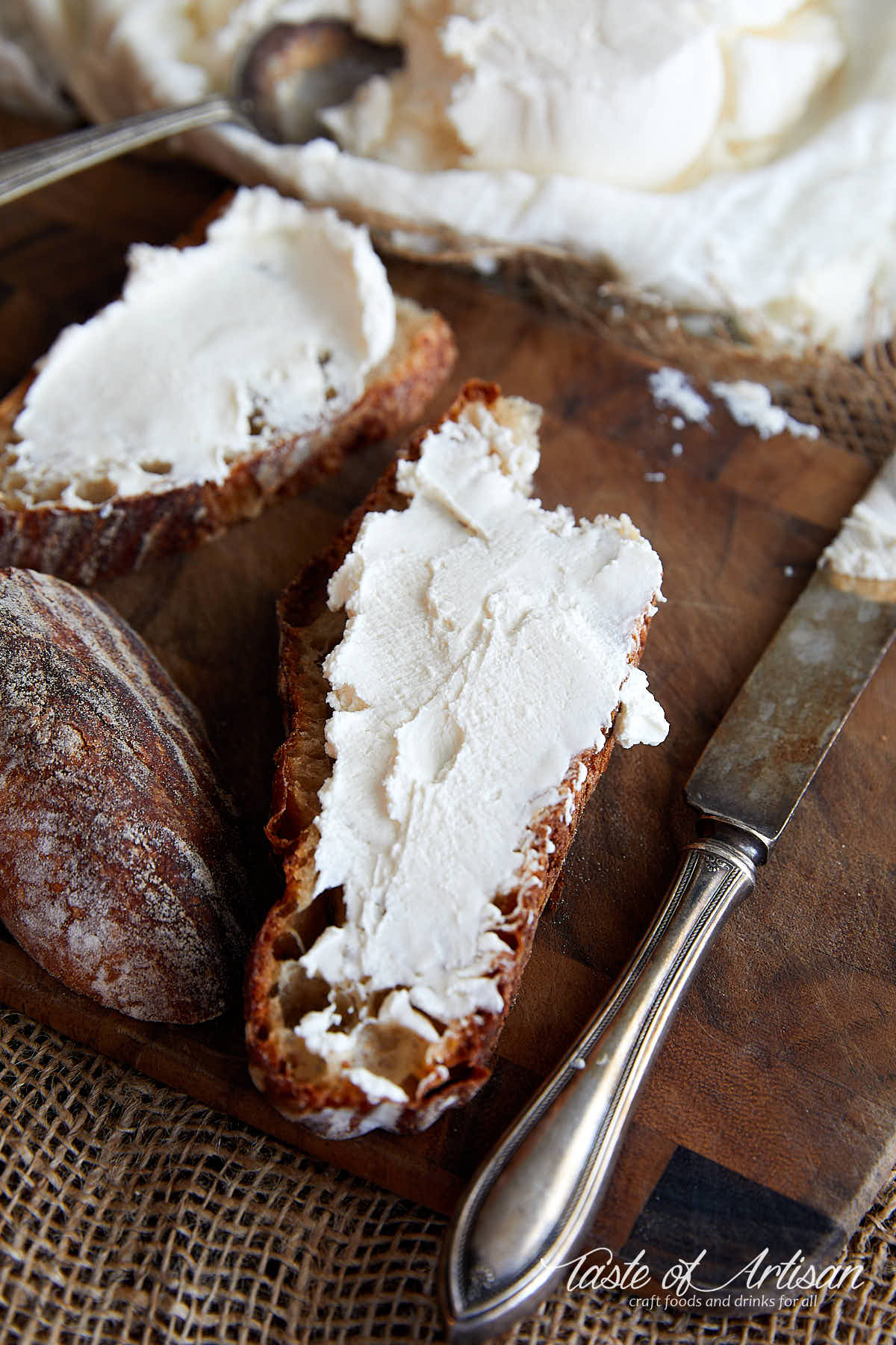 Slices of sourdough bread smeared with cream cheese on a cutting board, a bowl with cream cheese on the background.