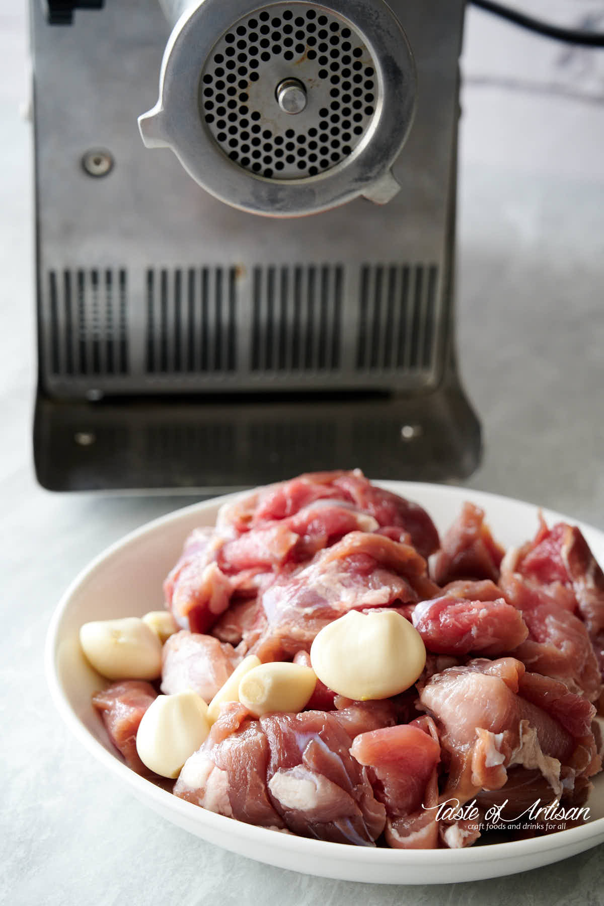 Less lean pork with garlic cloves in a bowl next to meat grinder.