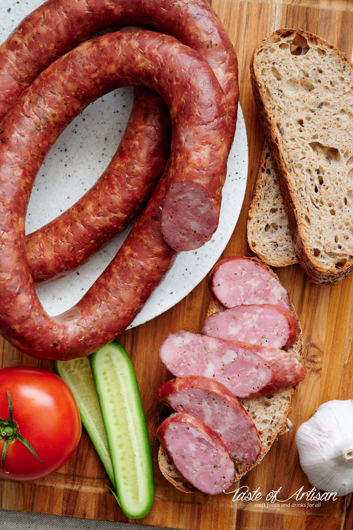 Whole and pieces of sliced garlic sausage on a table, with sliced bread and vegetables.