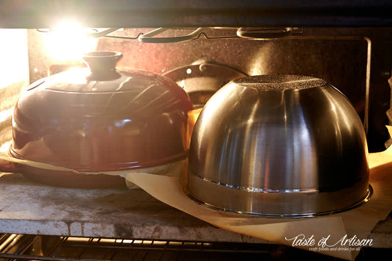 Two loaves in oven, one in a cloche, one under stainless bowl.