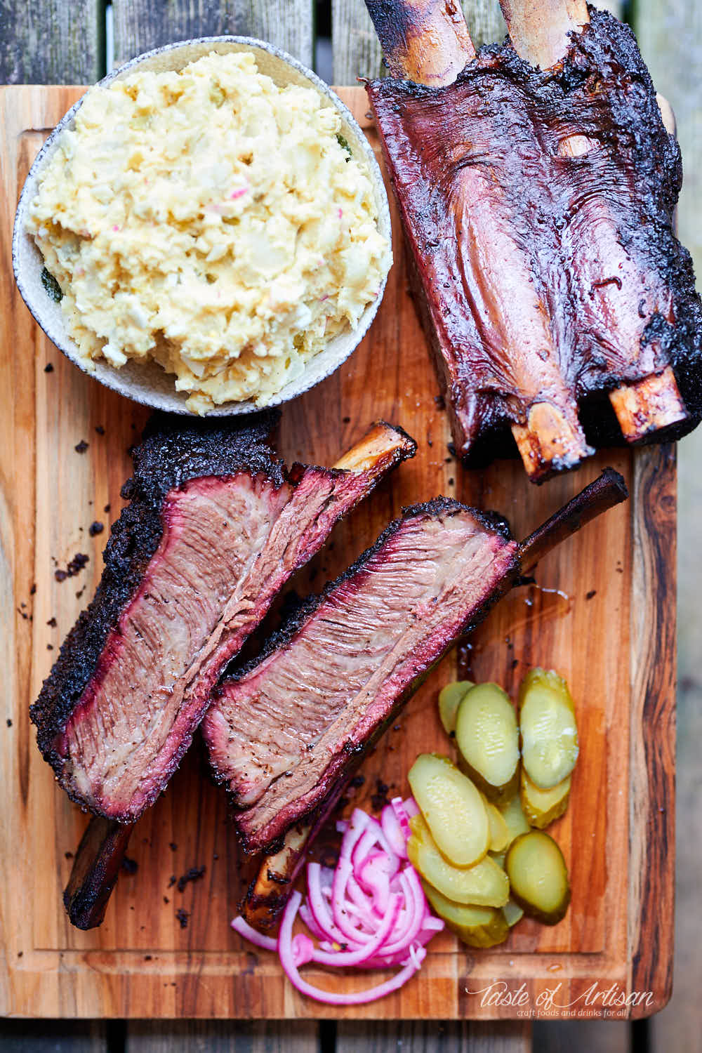 Sliced beef short ribs on a serving board with potato salad, pickles and pickled red onions.