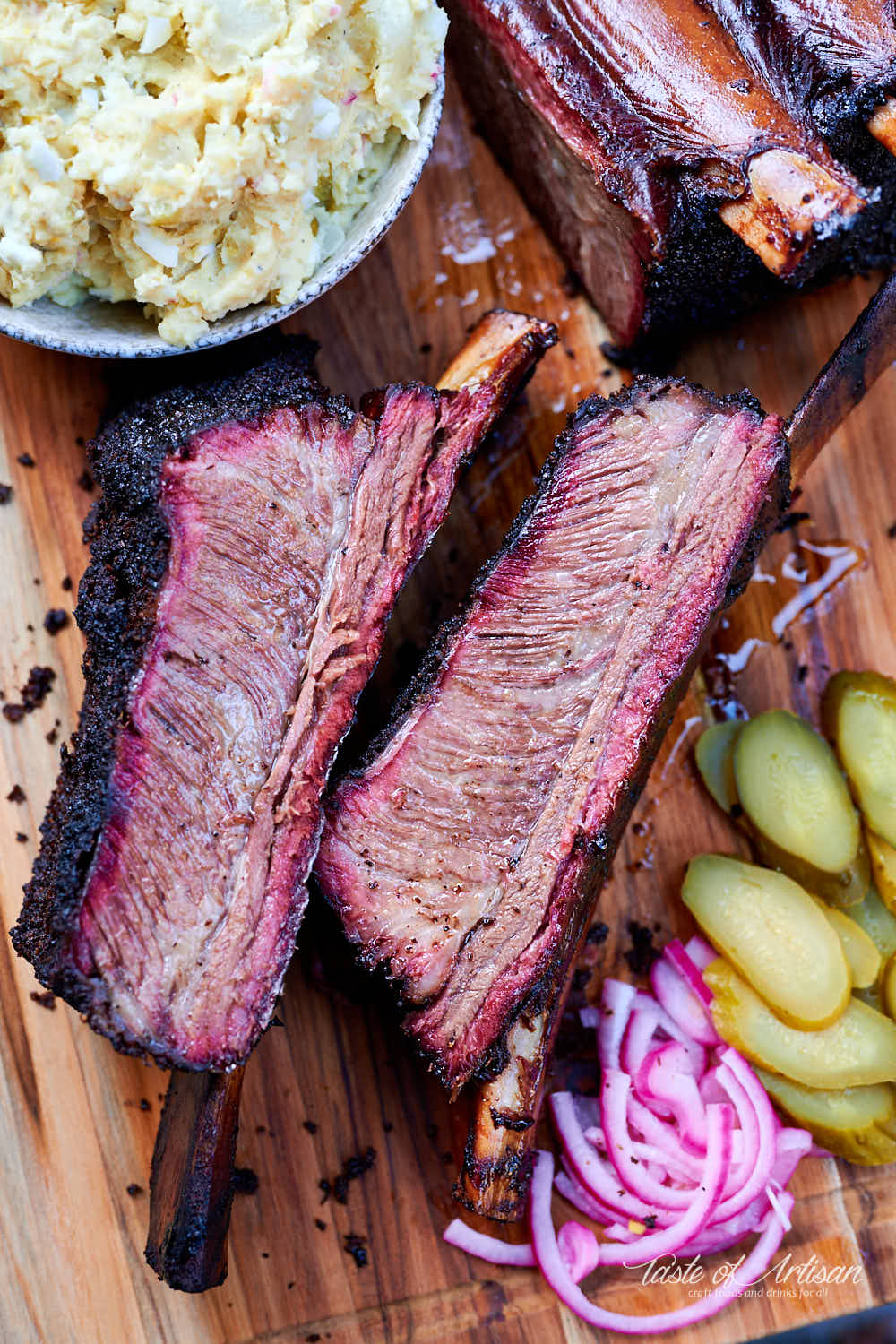 Beef short ribs on a serving board with potato salad, pickles and pickled red onions.